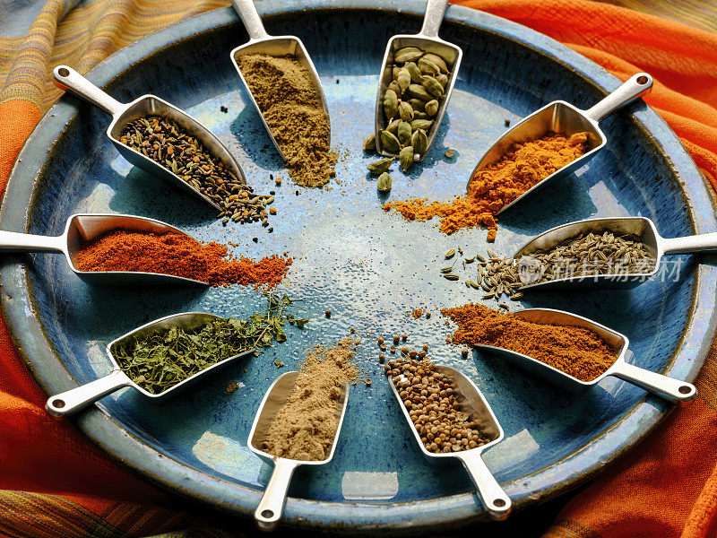 各种彩色的，有机的，干燥的，充满活力的印度食物香料，在金属测量的干燥食物勺在一个古老的蓝色/绿松石色的陶瓷盘子上，放在彩色的对比鲜明的桌布上。